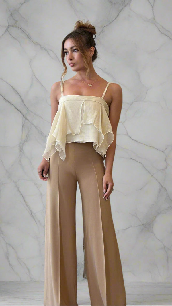 Woman wearing a beige top and pants standing in front of a white curtain.