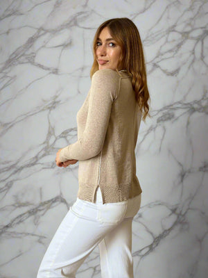 Woman wearing a beige sweater and white pants in a room with curtains.